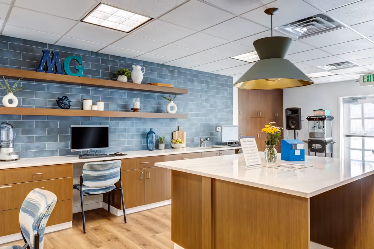 Interior view of a modern common area with a large kitchen island, wooden cabinets, blue tiled backsplash, two computer workstations with chairs, decorative shelves with plants and candles, a large pendant light, and a glass vase with yellow flowers on the island.
