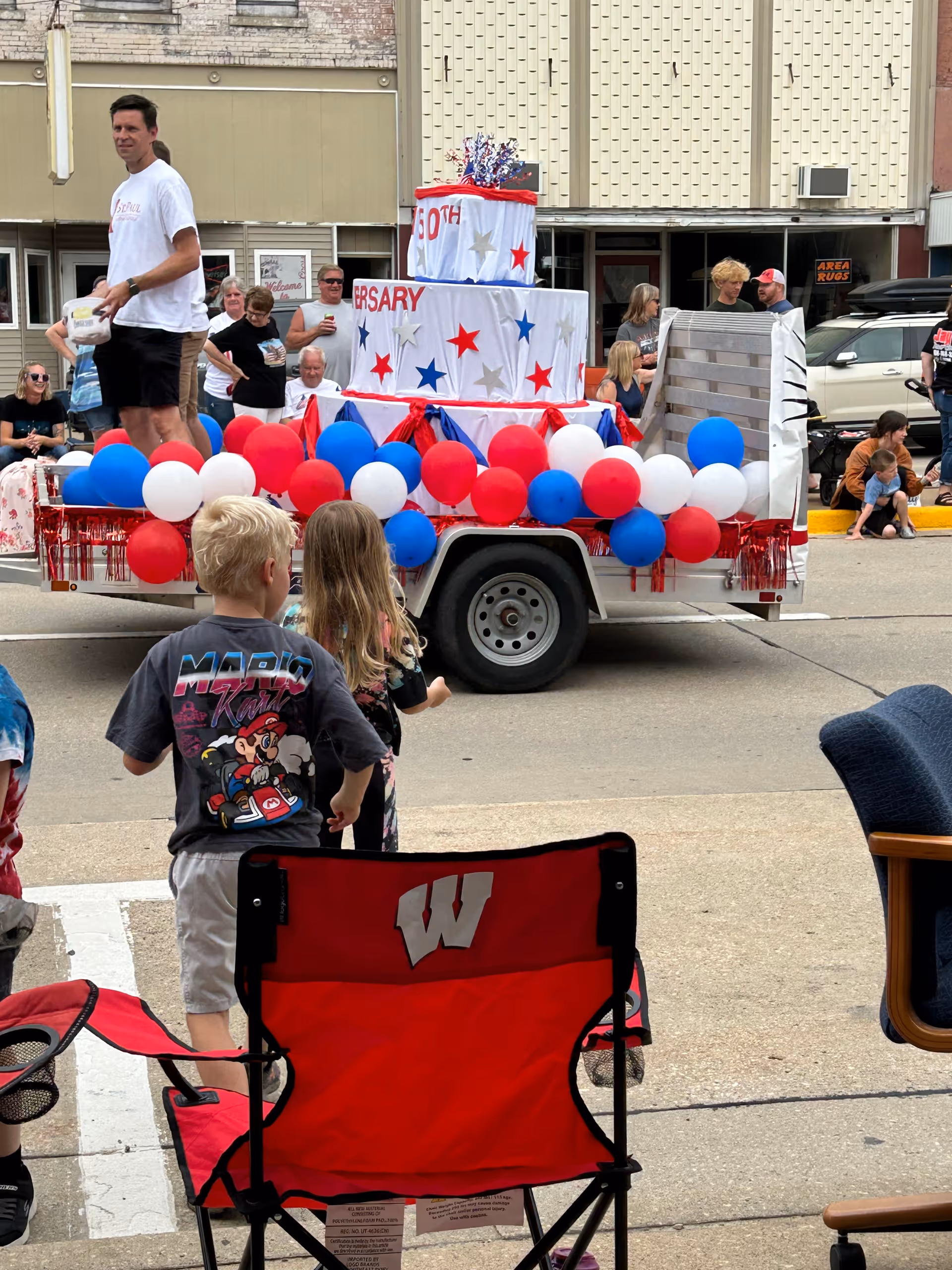 A parade float shaped like a tiered cake decorated with red, white, and blue balloons on a trailer as spectators, including children, watch on the street.