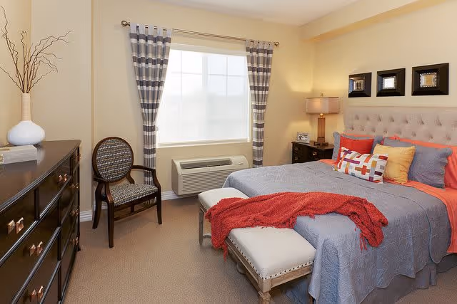 A cozy bedroom with a large bed covered in gray and orange bedding, multiple colorful pillows, and a red throw blanket. There is a cushioned bench at the foot of the bed, a wooden dresser with a white vase and decorative branches, a wooden chair with a patterned seat, a window with striped curtains, and a nightstand with a lamp and picture frame. The walls are painted light beige.