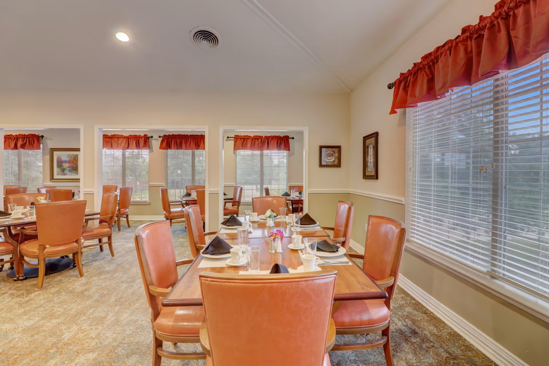 A dining room in a senior living facility with wooden tables set for meals, featuring cups, glasses, black napkins, and small flower vases. The room has multiple windows with red valances, beige walls, and carpeted floors. Several orange cushioned chairs surround the tables.
