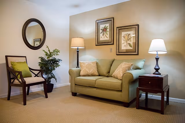 A cozy living room area with a green loveseat adorned with two patterned pillows, a wooden side table with a lamp, a wooden chair with a green cushion, a potted plant, a round wall mirror, and two framed botanical prints on the beige wall.