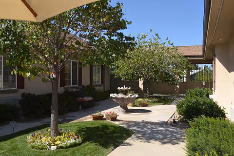 Outdoor courtyard area at Alta Ridge Memory Care of Sandy featuring a concrete walkway, green grass, flower beds, a tree with a birdhouse, a multi-tiered stone fountain, and surrounding bushes and plants under a clear blue sky.