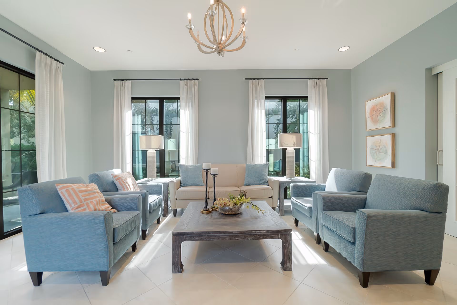 Bright living room with a sofa and four blue armchairs arranged around a wooden coffee table under a chandelier and large windows.