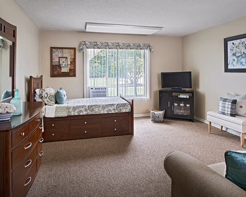 Bright furnished studio bedroom with a daybed, dresser, TV console and a large window with vertical blinds.
