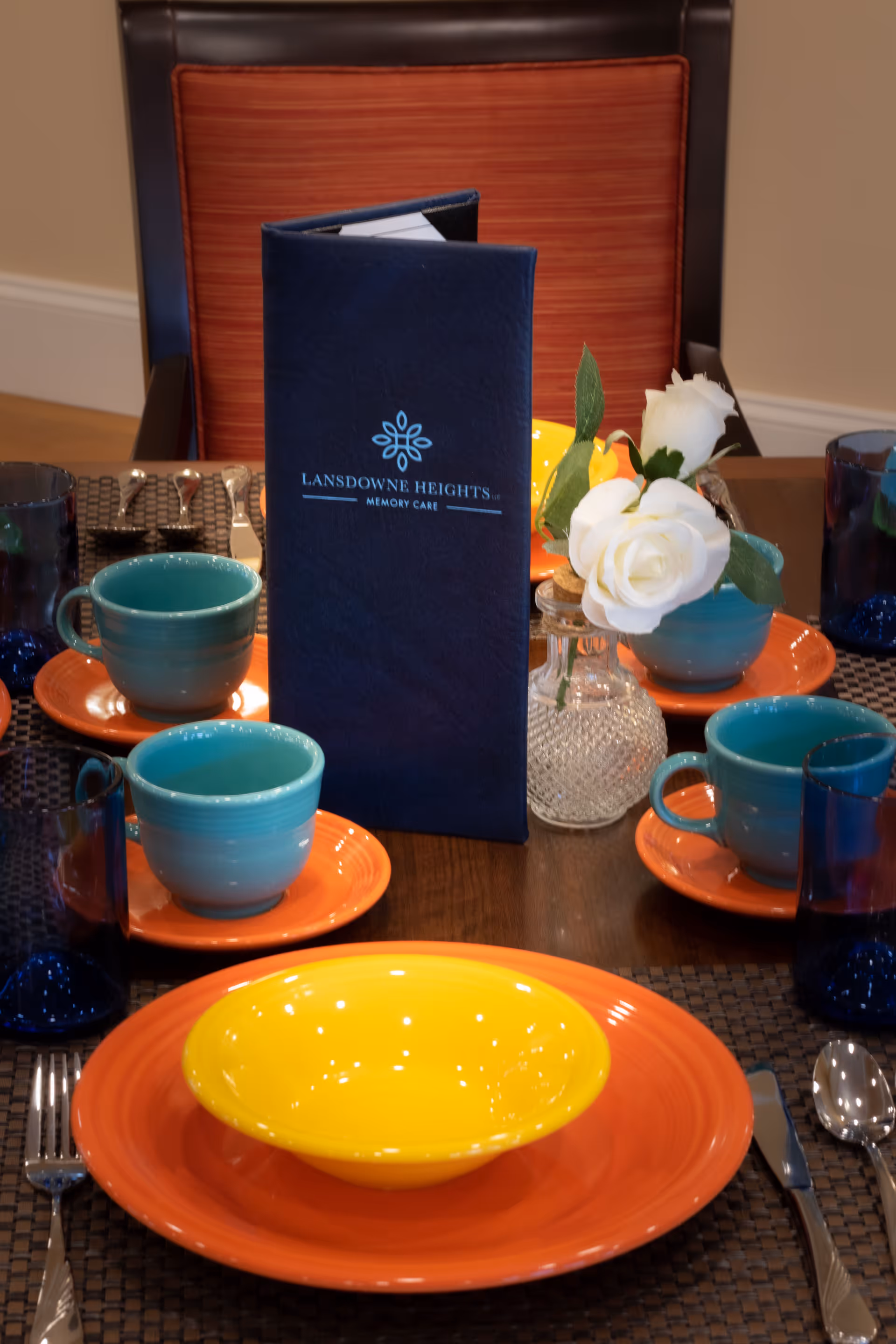 A dining table set with colorful orange plates, yellow bowls, turquoise cups, and blue glasses. A navy blue menu with the text 'Lansdowne Heights Memory Care' stands upright in the center. There is also a small vase with white flowers on the table, and a chair with a red cushion is visible in the background.