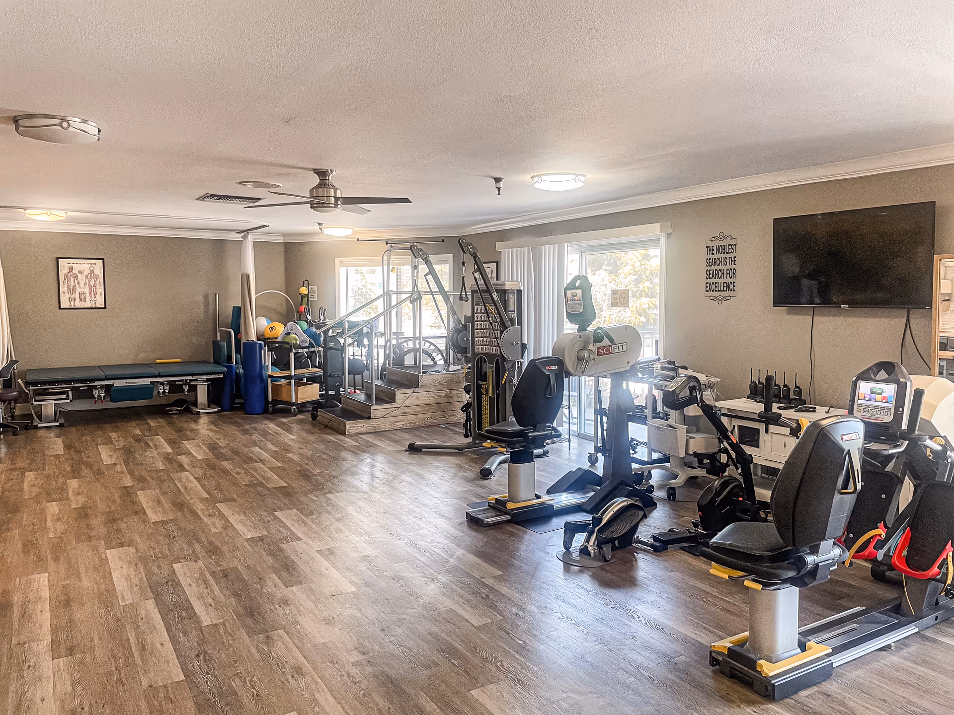 A spacious rehabilitation or physical therapy room with exercise equipment including stationary bikes, parallel bars, therapy mats, and various fitness tools. The room has wood-patterned flooring, neutral-colored walls, ceiling fans, and large windows letting in natural light. A flat-screen TV is mounted on the wall with a motivational quote beside it.