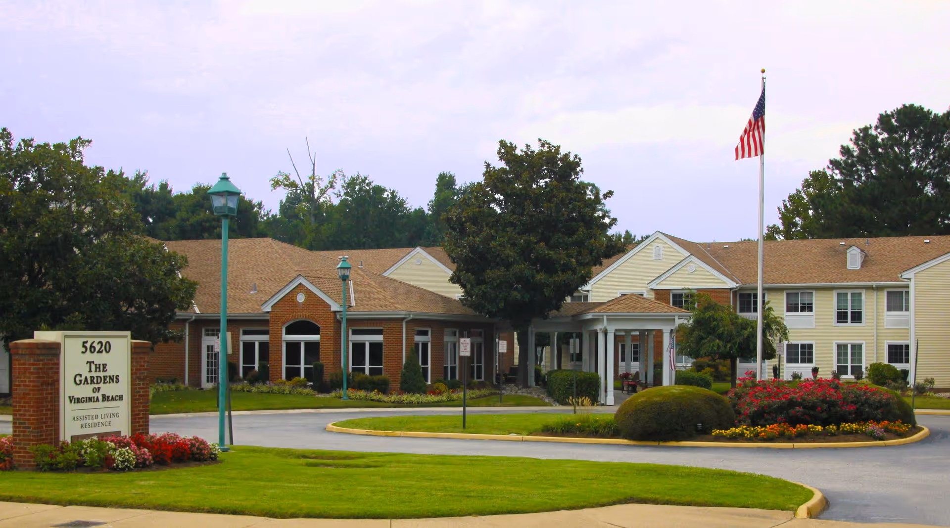 Exterior view of The Gardens of Virginia Beach assisted living residence showing a two-story building with beige siding and brick accents, a circular driveway with landscaped flower beds, an American flag on a flagpole, and green trees and shrubs surrounding the property.