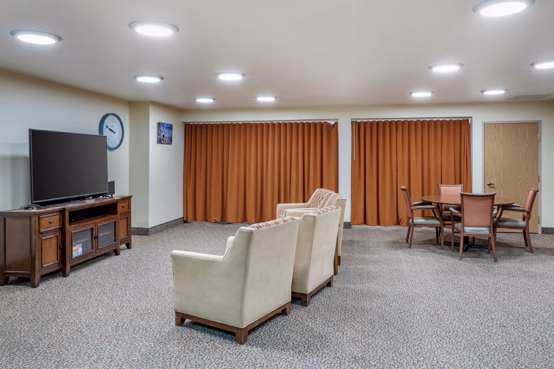 A spacious room with beige carpet and cream-colored walls featuring two beige armchairs facing a large flat-screen TV on a wooden TV stand. There is a round wooden table with four chairs in the corner, and two large windows covered with rust-colored curtains. A wall clock and a small framed picture are visible on the walls.