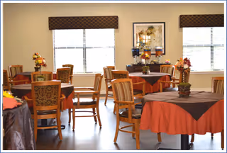 A dining room with multiple round tables covered with tablecloths in shades of brown and orange. Each table has a floral centerpiece. The room has large windows with brown valances, and framed artwork on the wall between the windows.