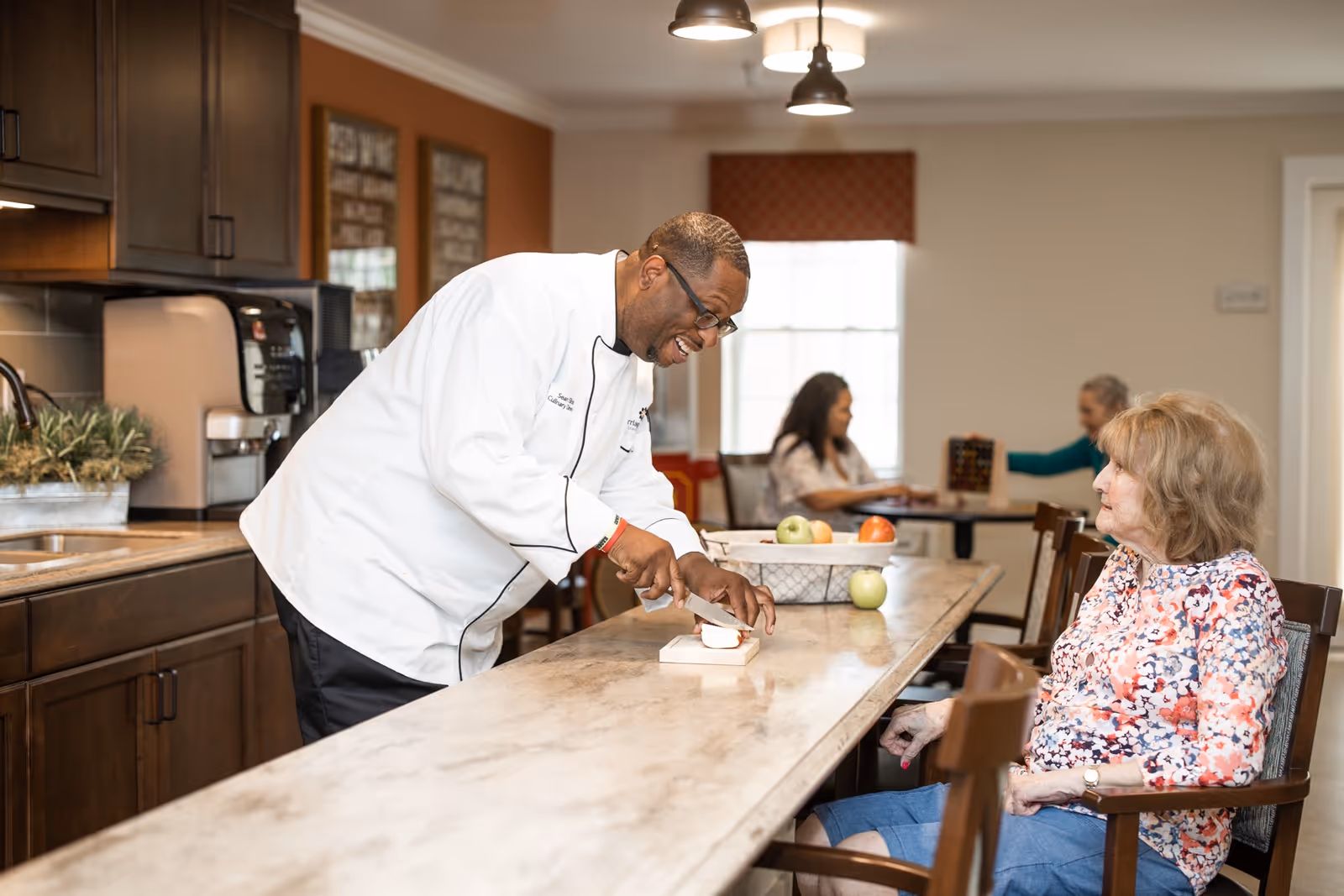 A chef in a white coat is slicing an apple on a kitchen counter while an elderly woman in a floral blouse and blue pants sits nearby watching. In the background, two other elderly women are seated at a table engaged in an activity. The setting appears to be a communal dining or kitchen area in a senior living facility.