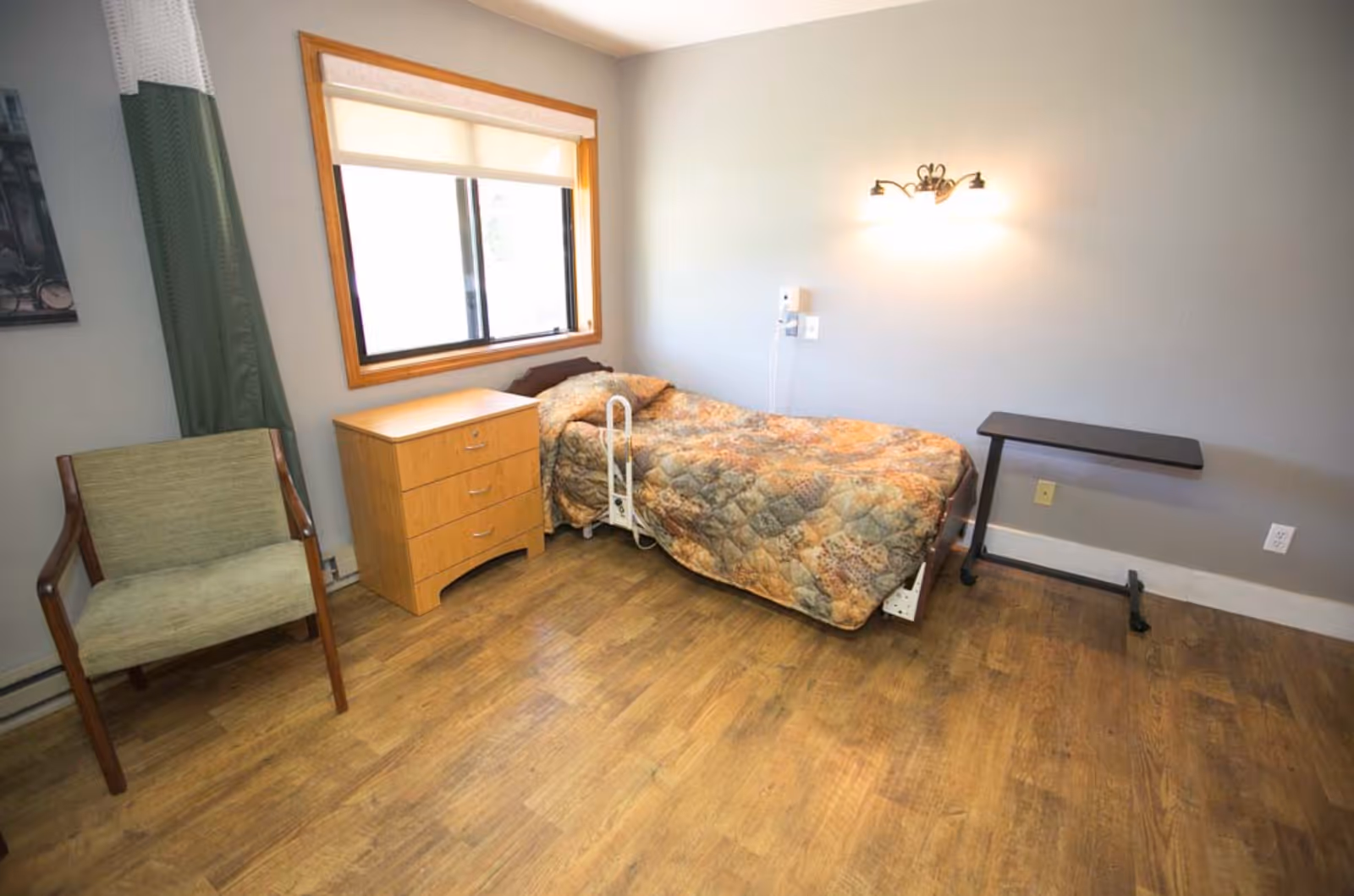 A small bedroom with a single bed covered in a patterned quilt, a wooden nightstand with drawers, a green upholstered chair, a window with a roller shade, a wall-mounted light fixture, and a small adjustable overbed table on wheels. The floor is wood and the walls are painted light gray.
