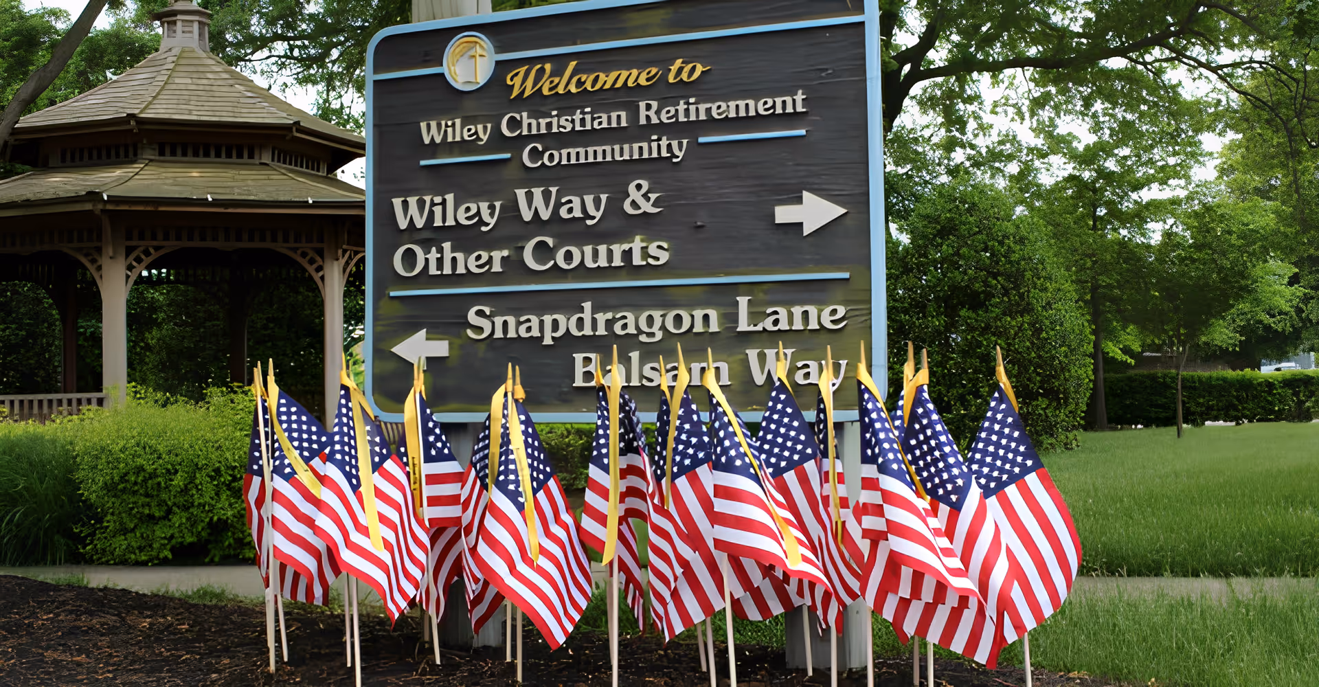 Entrance sign for Wiley Christian Retirement Community with a row of American flags and a gazebo in the background.