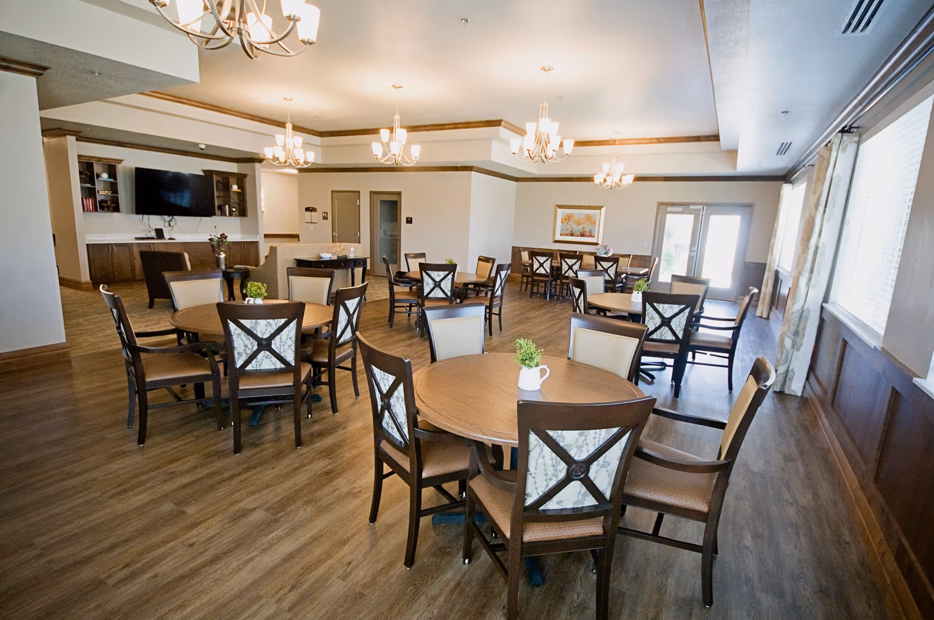 A spacious dining room with multiple round wooden tables and chairs arranged neatly. The room features wooden flooring, large windows with curtains allowing natural light, chandeliers hanging from the ceiling, and a wall-mounted TV with cabinetry underneath.