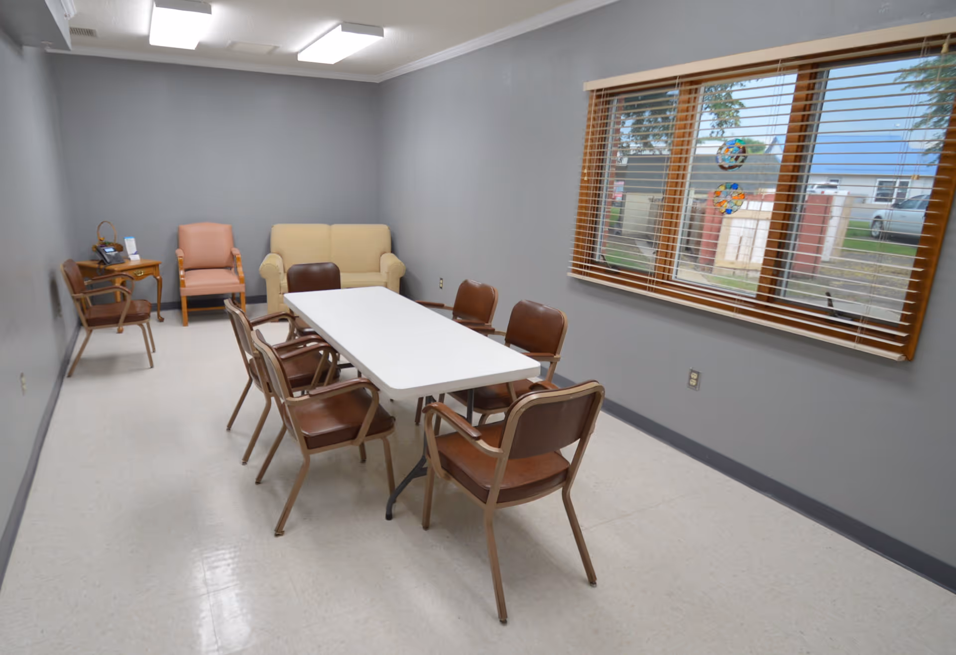 A small meeting or dining room with a white rectangular table surrounded by six brown chairs. Against the far wall, there is a beige loveseat and two additional chairs, one pink and one brown. A wooden side table with a phone and some papers is placed in the corner. The room has gray walls, a large window with wooden blinds, and a view of the outside.