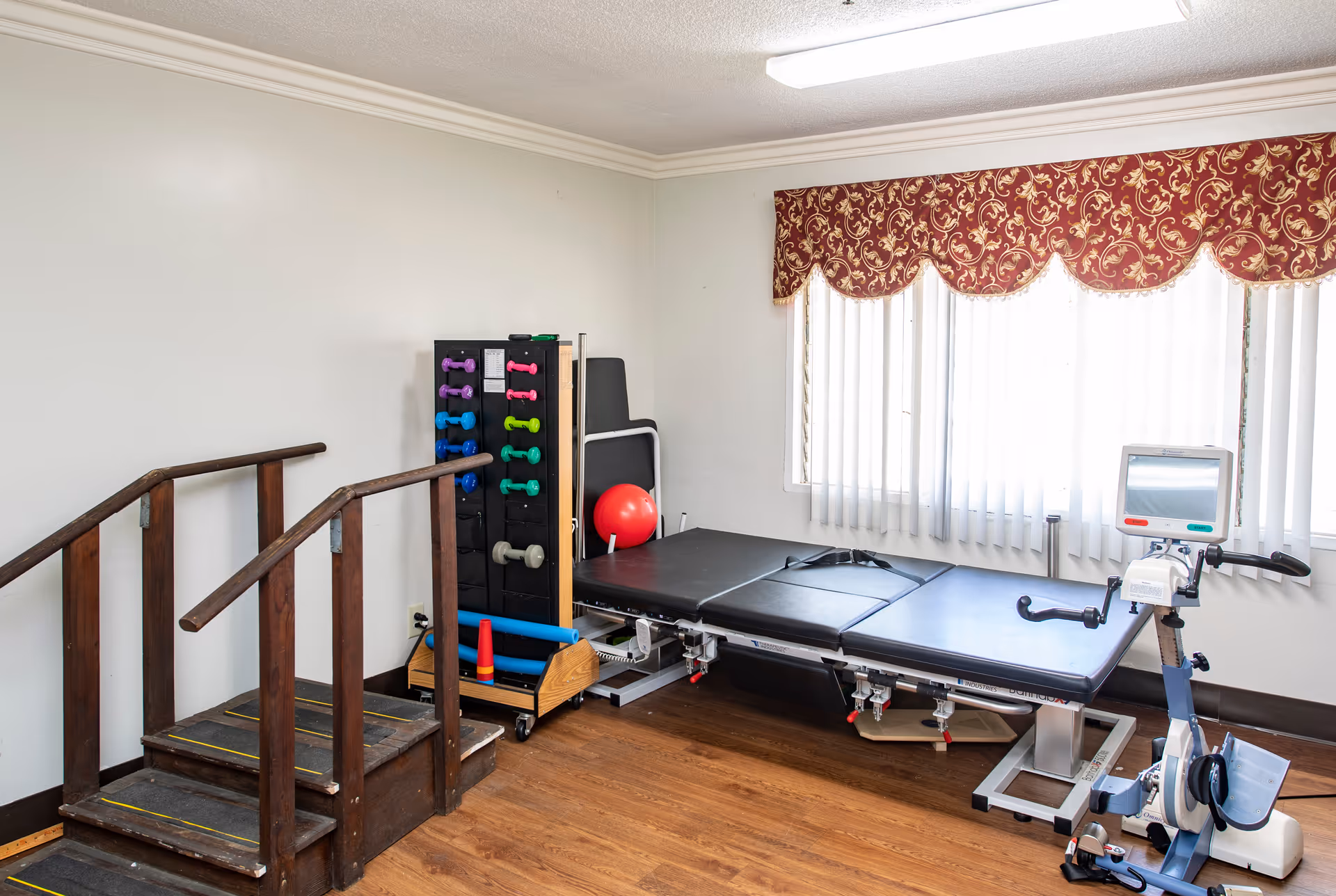 A physical therapy room with wooden stairs for rehabilitation, a rack of colorful dumbbells, a red exercise ball, a black padded therapy table, and a stationary exercise bike near a window with vertical blinds and a red patterned valance.