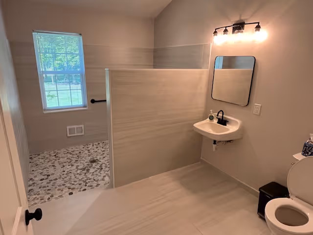 Bright accessible bathroom with a curbless pebble-floor walk-in shower, half wall, wall-mounted sink with mirror, toilet, and a window.