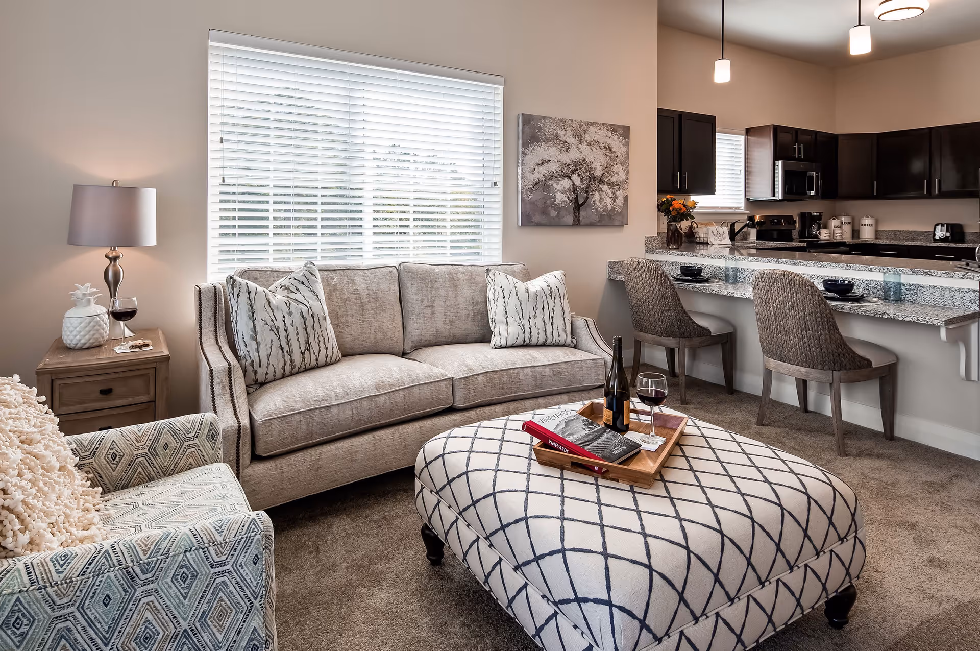 Bright living room with a beige sofa, patterned ottoman, armchair, and an open kitchen with bar seating.