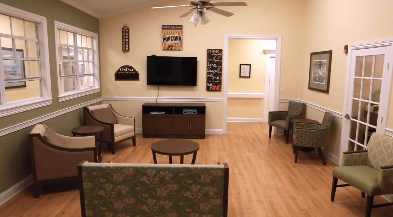 A cozy living room area with multiple armchairs and a loveseat arranged around two small round tables. A flat-screen TV is mounted on the wall above a wooden cabinet. The walls are painted in light yellow and green tones, decorated with framed pictures and signs including one that says 'Popcorn' and another that says 'Cinema Always Showing'. There is a ceiling fan with lights above and a doorway leading to another room.