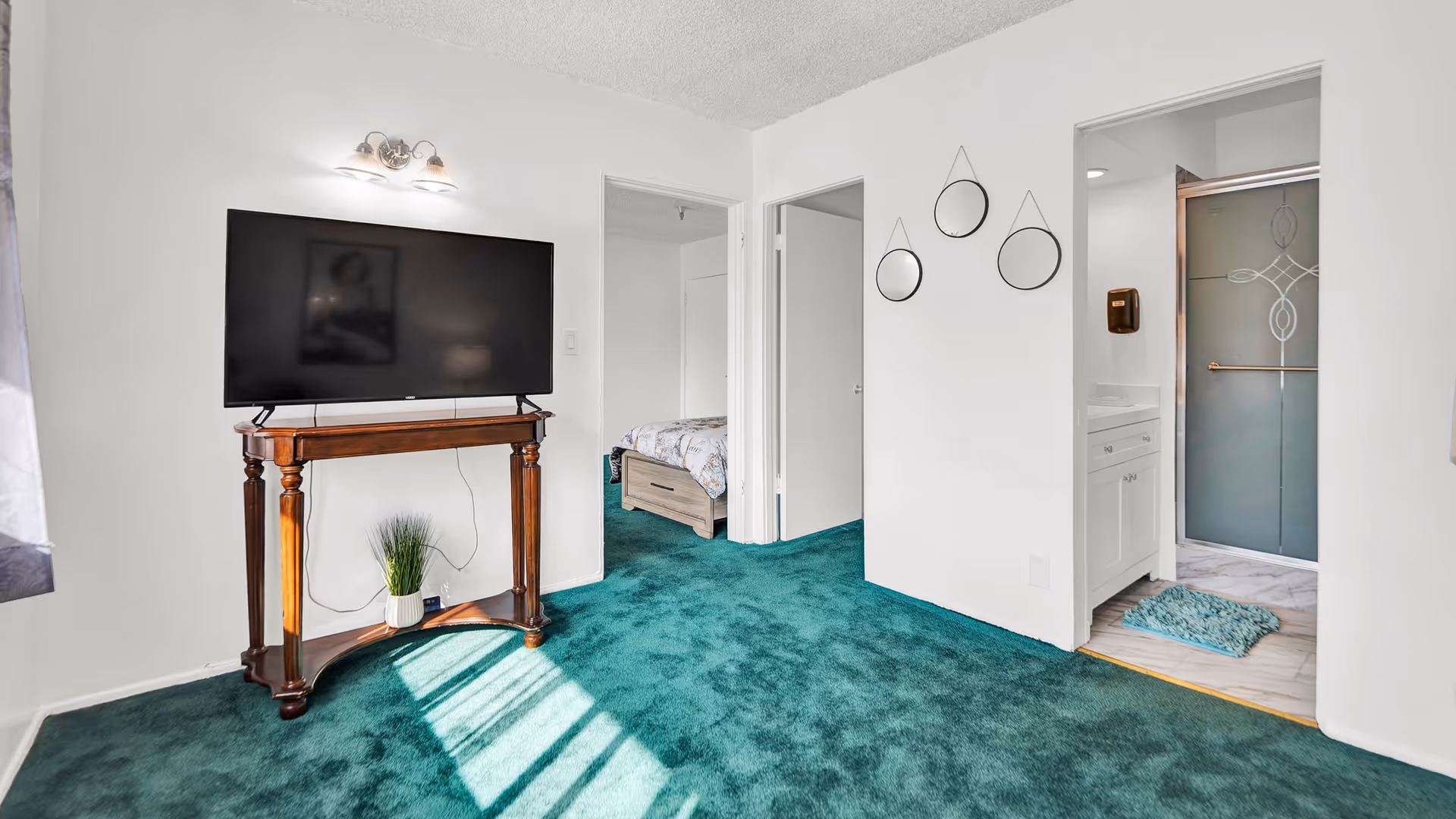 Interior living area with green carpet, a large TV on a wooden stand, and open doorways leading to a bedroom and a bathroom.