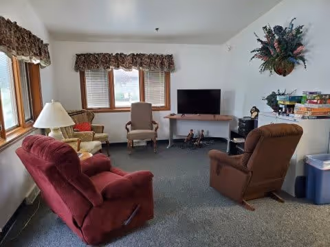 A cozy living room with a red recliner, a brown recliner, and two wooden chairs arranged around a TV on a small table. The room has carpeted floors, windows with floral valances, a lamp on a side table, a decorative wall hanging with flowers, and a small shelf with board games and other items.