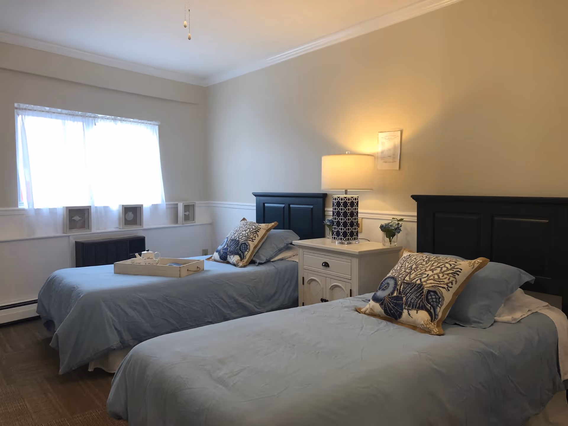 A bedroom with two twin beds featuring blue bedding and decorative pillows with seashell designs. Between the beds is a white nightstand with a blue and white patterned lamp and a small vase with flowers. A window with sheer white curtains lets in natural light, and there are small framed pictures on the windowsill.