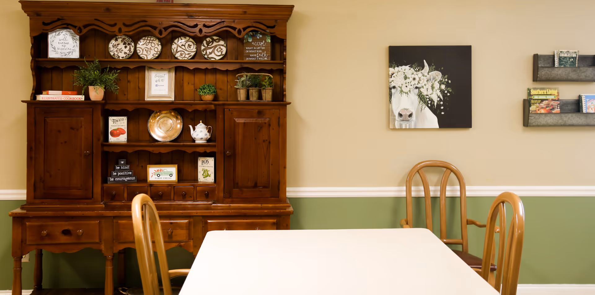 Dining area with a white table, wooden chairs and a large wooden hutch decorated with plates, plants, and artwork on a green-beige wall.