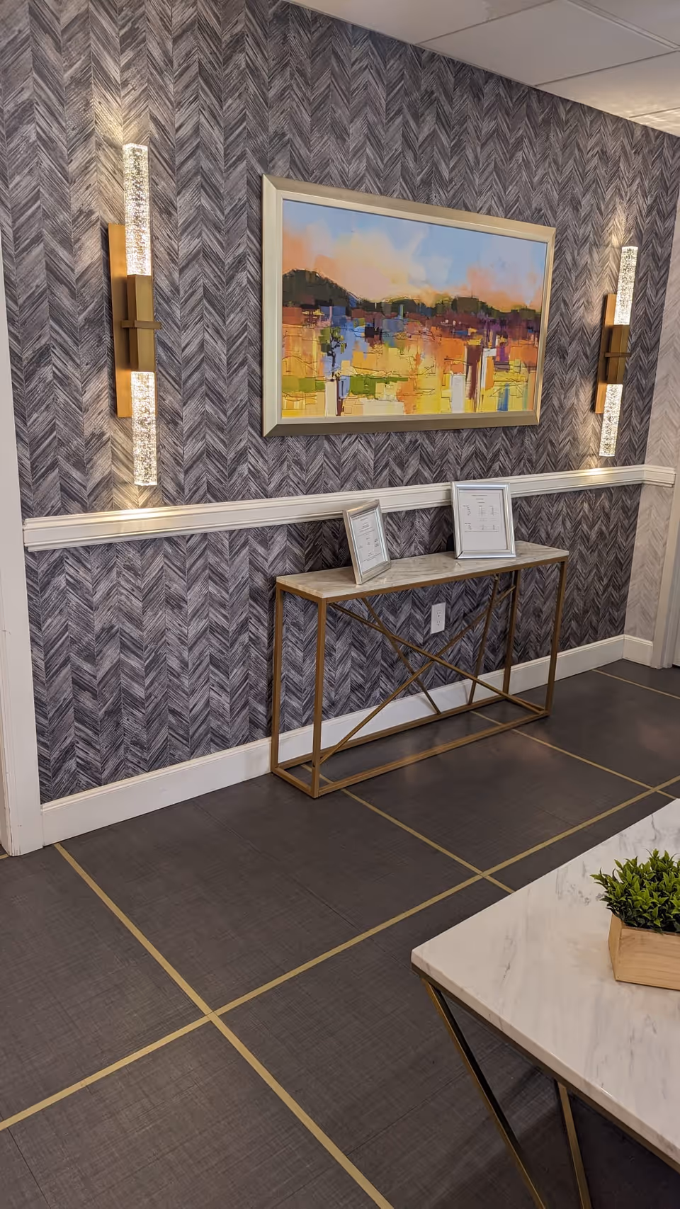 Interior wall with textured gray chevron wallpaper, two vertical modern wall sconces with crystal-like detailing, a framed abstract painting with colorful brush strokes, and a narrow console table with a gold metal frame and a marble top holding two small framed pictures. The floor has large dark tiles with gold grid lines, and a corner of a marble-topped table with a small potted plant is visible.