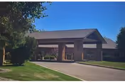 Exterior view of a single-story brick building with a covered entrance driveway, surrounded by green grass and trees under a clear blue sky.