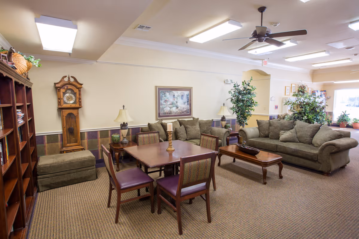 A cozy living room area in Wesley House featuring two green upholstered sofas, a wooden coffee table, a square wooden table with four chairs, a grandfather clock, two table lamps, a large bookshelf filled with books, and several potted plants. The room has beige walls, carpeted flooring, and ceiling lights with a ceiling fan.