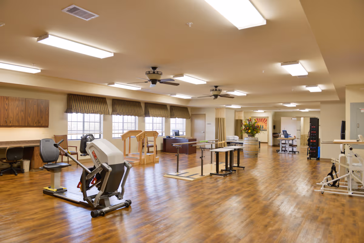 A spacious rehabilitation or physical therapy room with wooden flooring, exercise equipment including a seated rowing machine, parallel bars for walking practice, and other therapy apparatus. The room has multiple windows with striped valances, ceiling fans, and bright overhead lighting. There is a desk and chair along one wall and a large floral arrangement on a barrel in the background.