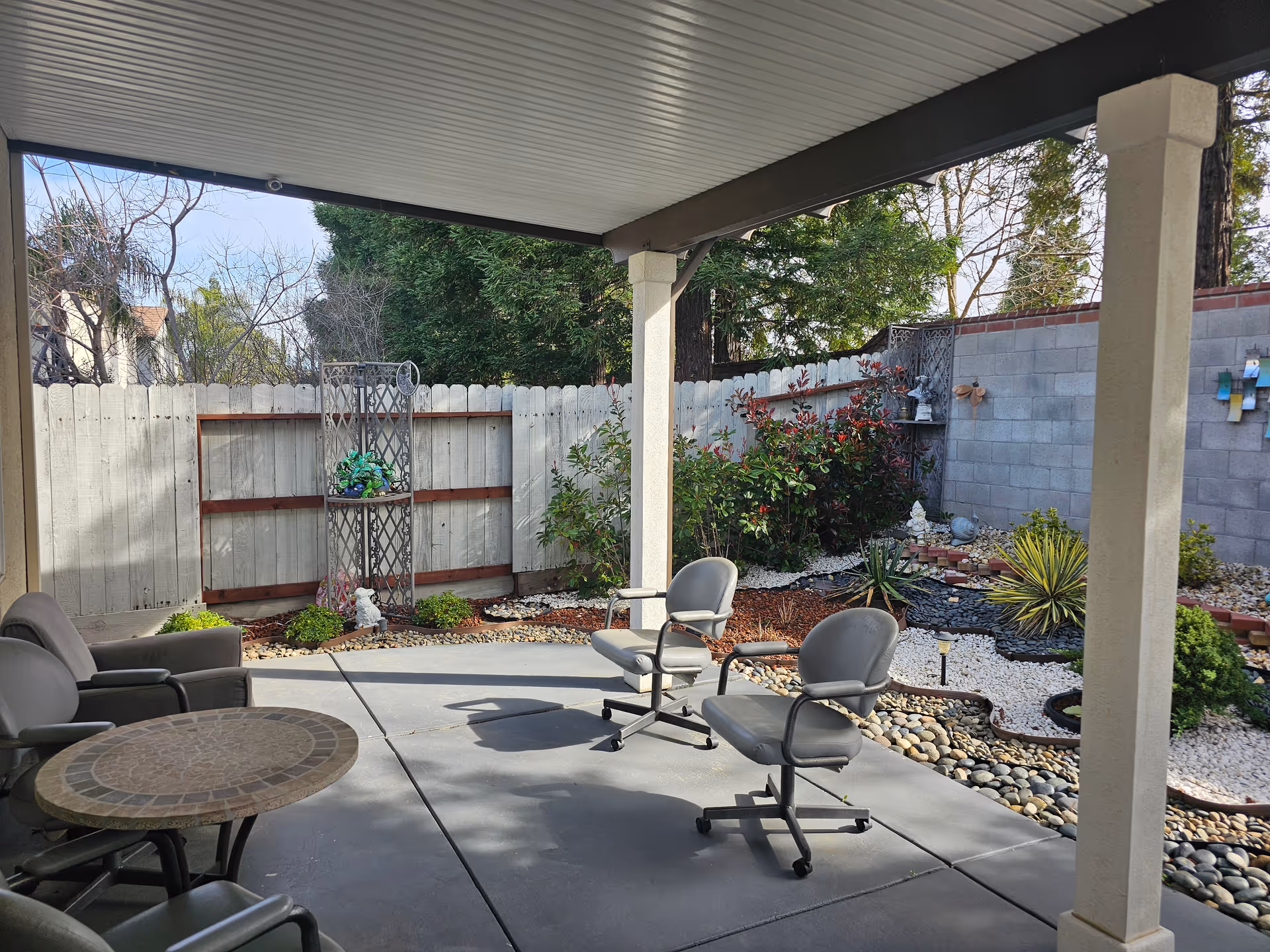 Covered patio area with a round mosaic table and several chairs on a concrete floor. The patio overlooks a landscaped garden with various plants, shrubs, and decorative rocks, enclosed by a wooden fence and a cinder block wall.