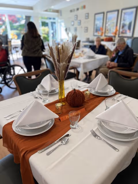 A dining table set for four with white plates, folded white napkins, silverware, and glasses. The table has a white tablecloth with an orange runner, decorated with a small knitted pumpkin, two small candle holders, and a yellow vase holding dried pampas grass. In the background, elderly people are seated at another table in a bright room with large windows and framed pictures on the wall.