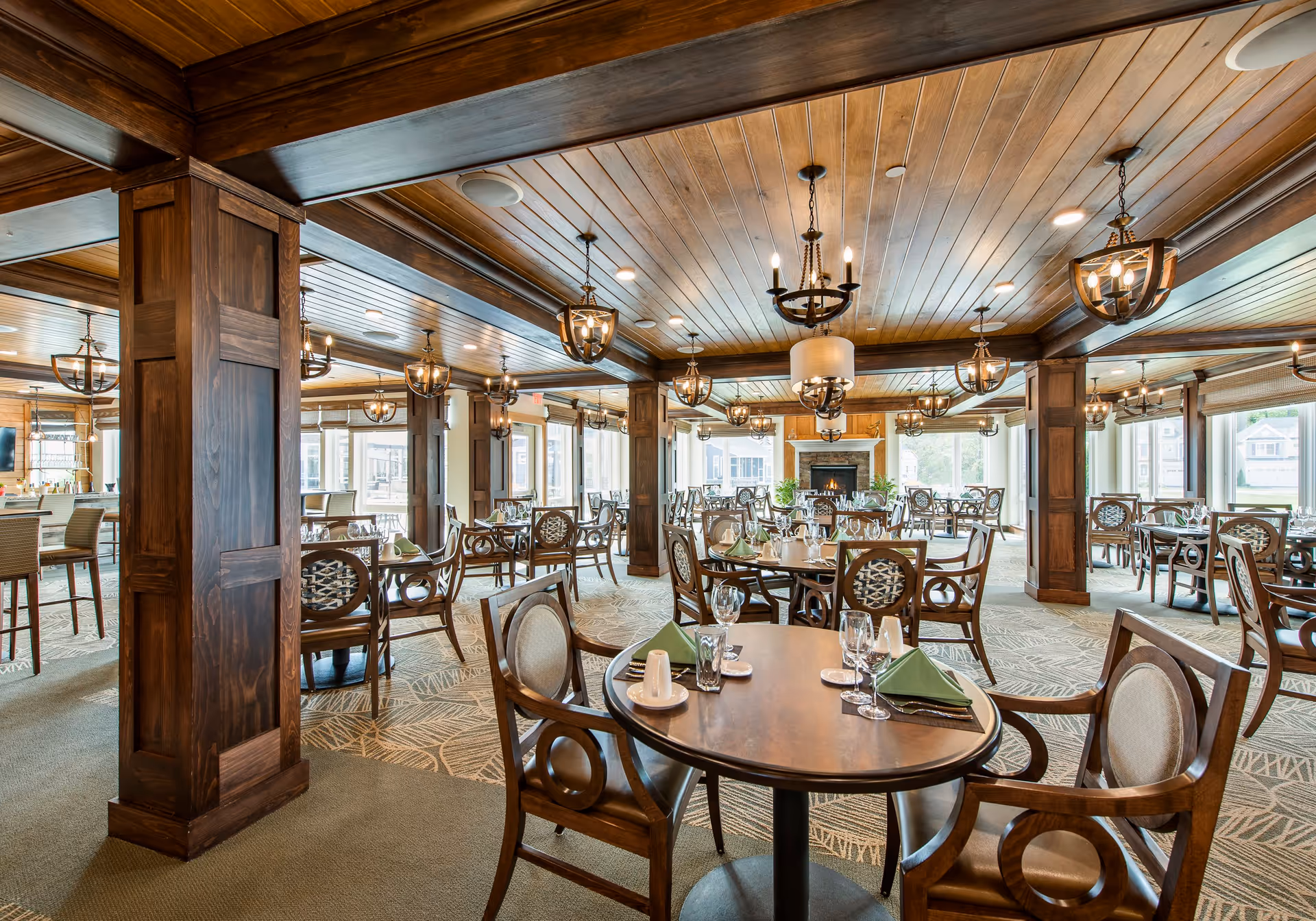 A spacious dining room with wooden ceiling and pillars, featuring multiple round tables set with glassware, napkins, and silverware. The room is well-lit with hanging chandeliers and large windows allowing natural light. A fireplace is visible at the far end of the room.