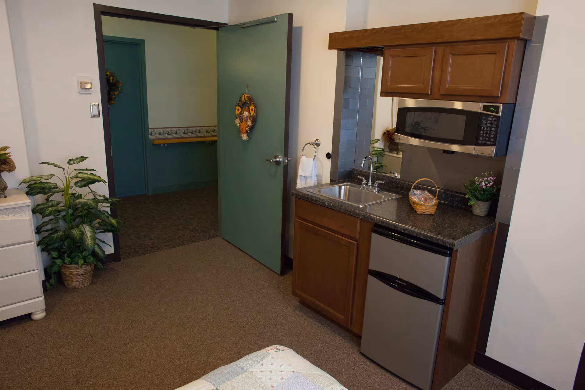 Interior view of a senior living facility room showing a small kitchenette with a sink, microwave, mini refrigerator, and wooden cabinets. There is an open green door decorated with a wreath, leading to a hallway with green walls and a handrail. A potted plant and a white dresser are visible on the left side.