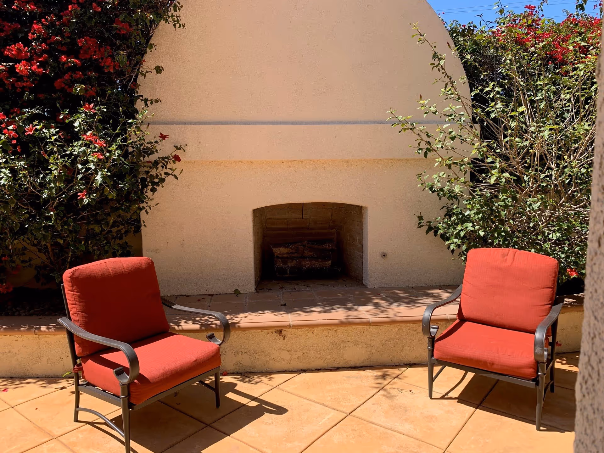 Outdoor patio area with two red cushioned chairs facing a built-in outdoor fireplace surrounded by plants and flowers under a clear blue sky.
