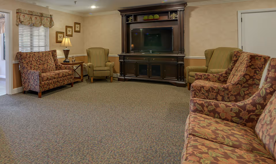 A cozy living room area with patterned armchairs and a loveseat arranged around a large dark wooden entertainment center with a flat-screen TV. The room has beige walls with a chair rail, carpeted floor, a side table with a lamp, and framed butterfly artwork on the wall.
