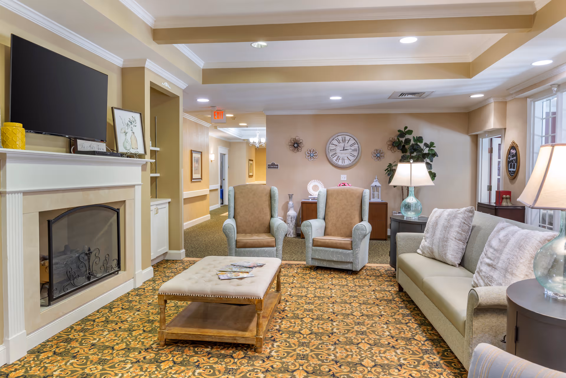 Communal lounge with sofa, armchairs, a fireplace and TV, coffee table, lamps, and a decorative wall clock.