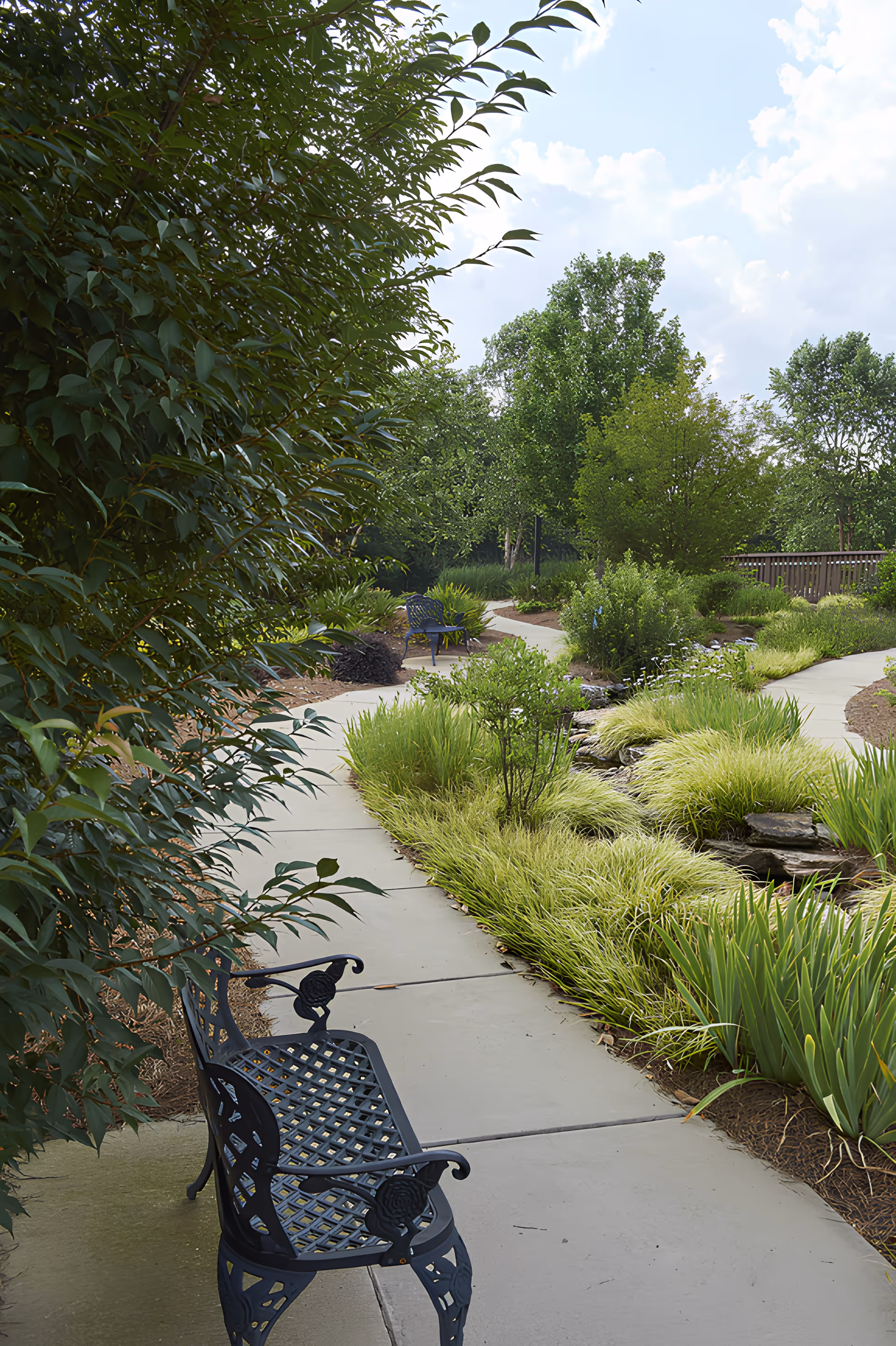 A winding concrete pathway through a lush garden with green shrubs, trees, and ornamental grasses. There are black metal benches along the path for seating, and the sky is partly cloudy.