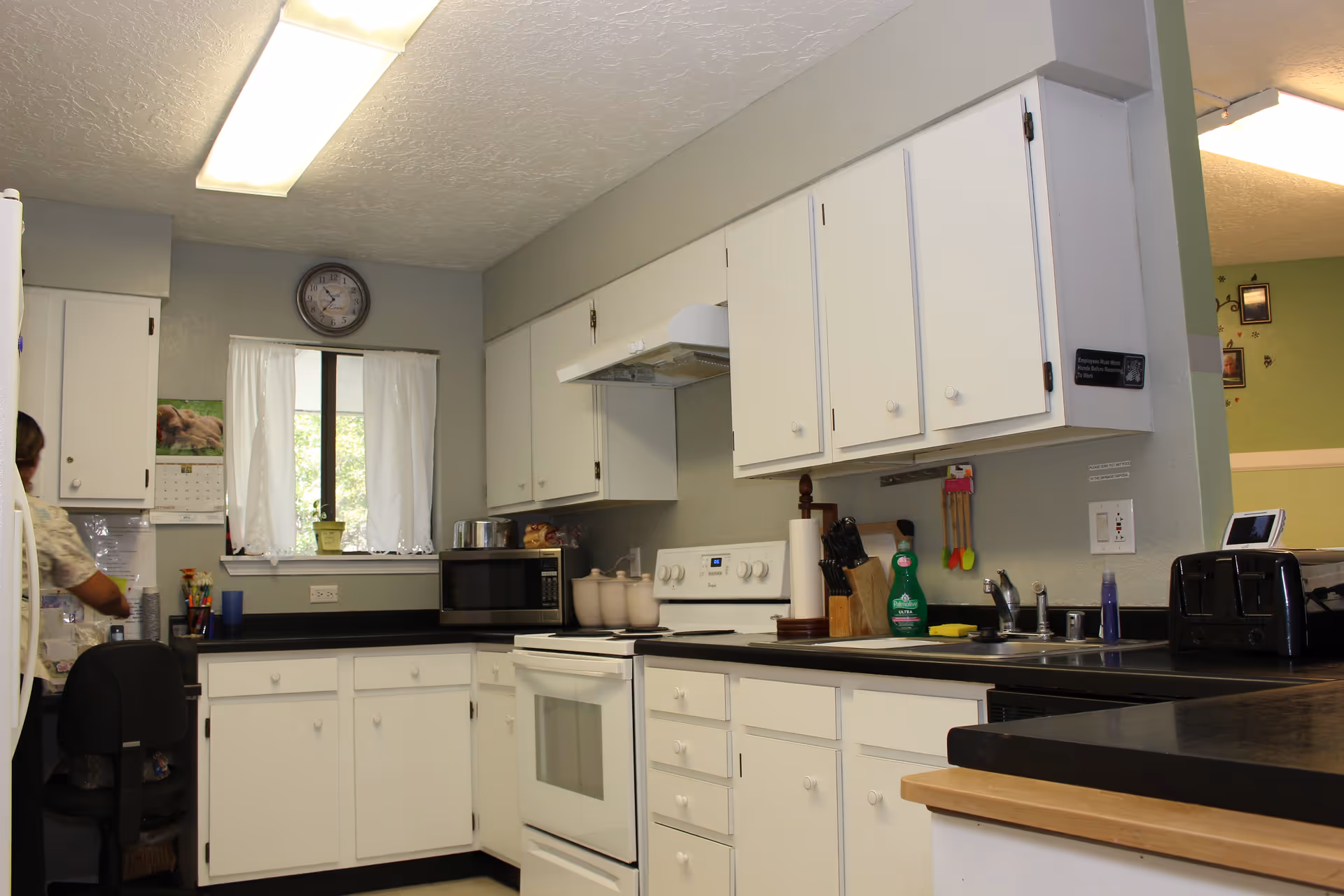 A clean kitchen with white cabinets and black countertops. The kitchen features a white stove with an overhead vent, a microwave, a toaster, a sink with dish soap, and a knife block. There is a window with white curtains and a clock above it. A person is partially visible on the left side near a refrigerator.