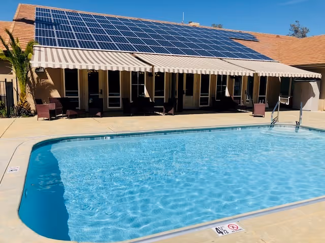 Outdoor swimming pool with lounge chairs and a building with striped awnings and solar panels on the roof.