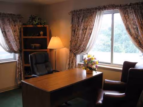 A small furnished office with desk, chairs, lamp, bookshelf, and a window.