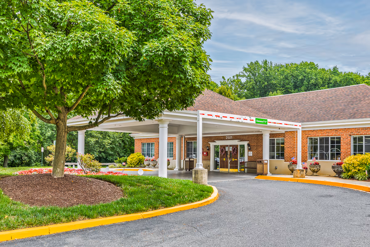 Covered entrance of a brick senior living facility with white columns, a circular driveway, landscaped flower beds, and mature trees.