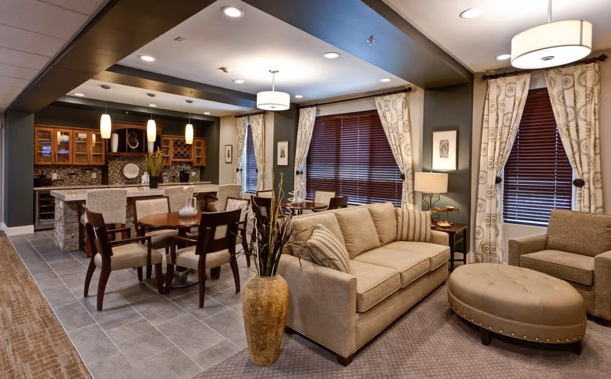 A cozy and well-lit living area in an assisted living facility featuring a beige sofa with striped pillows, a matching armchair, and a large oval ottoman. Behind the seating area, there are two round wooden tables each surrounded by four chairs. The space includes a kitchen area with a stone island, bar stools, hanging pendant lights, and wooden cabinets with glass doors. The windows are adorned with patterned curtains and wooden blinds, and the room has a warm, inviting atmosphere.