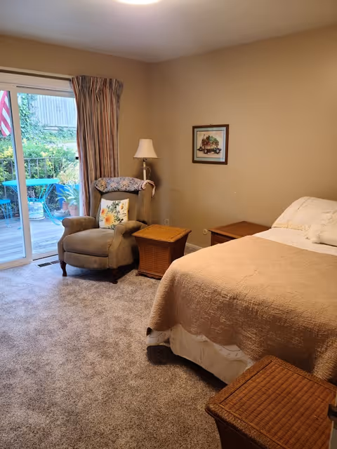A cozy bedroom with a beige bedspread on a bed, a cushioned armchair with a floral pillow and a throw draped over the back, two wicker side tables, a floor lamp, a framed picture on the wall, and sliding glass doors leading to an outdoor patio with a table and chairs surrounded by greenery.