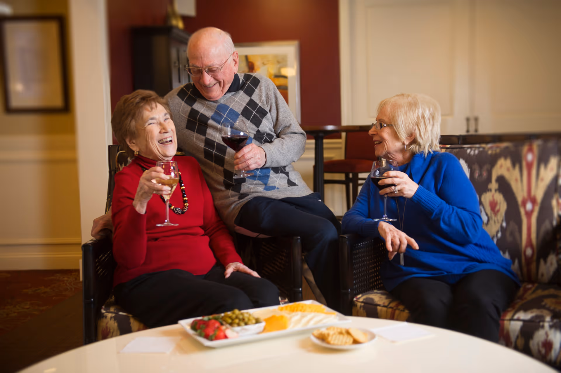Three elderly people sitting in a cozy living room area, smiling and holding glasses of wine. A man in a gray argyle sweater is sitting between two women, one in a red sweater and the other in a blue sweater. In front of them is a table with a plate of assorted snacks including cheese, crackers, olives, and strawberries.