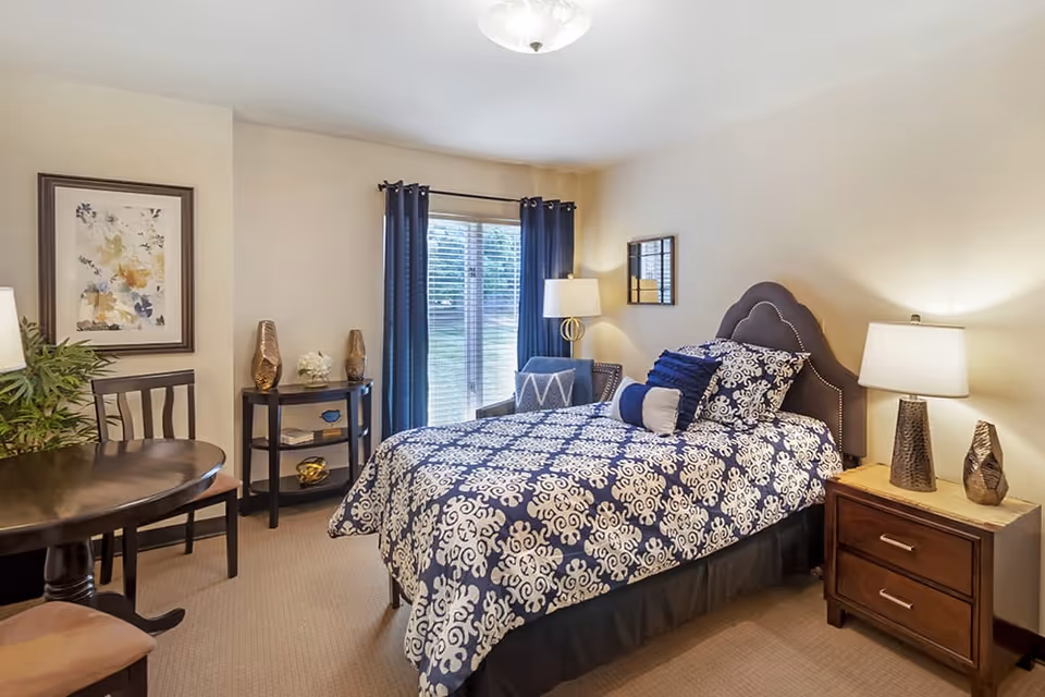 Well-lit bedroom with a patterned blue-and-white bed, nightstand and lamp, small table and chairs, and a window with dark curtains.
