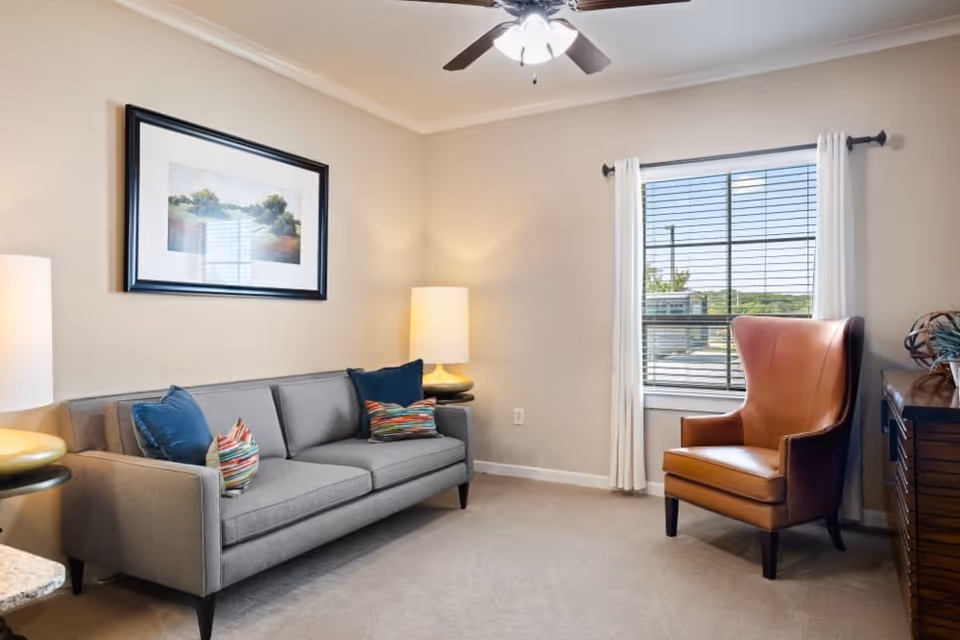 A cozy living room with a gray sofa adorned with colorful pillows, a brown leather armchair near a window with white curtains, a ceiling fan with lights, a framed landscape painting on the wall, and two table lamps providing warm lighting.