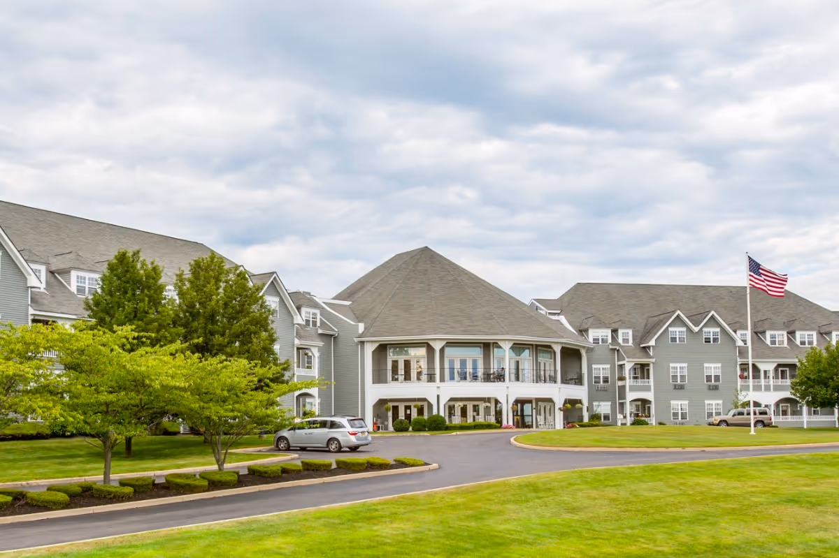 Exterior view of a large senior living facility building with gray siding and multiple windows. The building has a central section with a covered porch and balcony. There is a driveway with a parked silver vehicle, well-maintained green lawns, trees, and an American flag on a flagpole. The sky is partly cloudy.