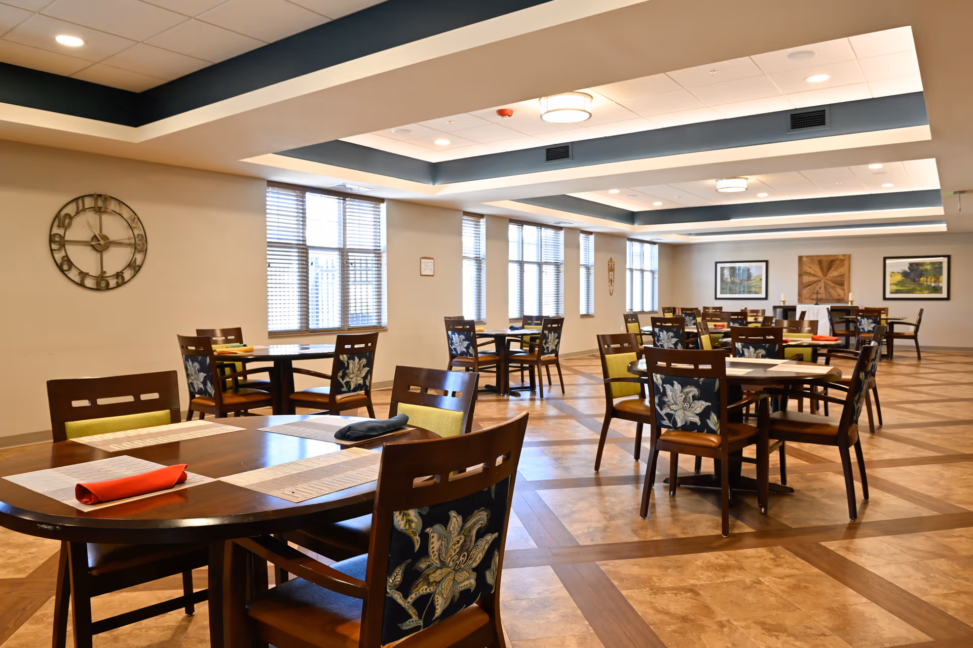 A spacious dining room with multiple wooden tables and chairs arranged neatly. The chairs have floral patterned backs and some have green cushions. The room has large windows with blinds allowing natural light to enter. The ceiling features recessed lighting and decorative beams. There are framed artworks on the far wall and a large decorative clock on the left wall.