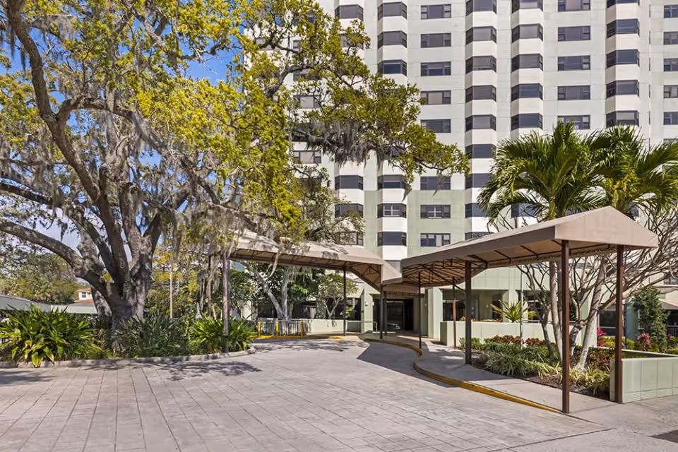 Entrance of a multi-story building with covered drive-up canopies, landscaped planters and large trees in front.